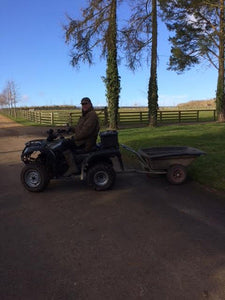 Man sat on ride on mower with wheelbarrow trailer attached